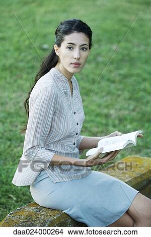 Picture - Woman sitting outdoors, reading book, looking up at camera. Fotosearch