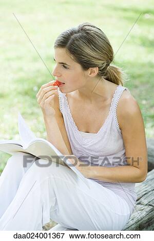 Stock Photo - Woman sitting outdoors, reading book and eating cherry tomatoes. Fotosearch