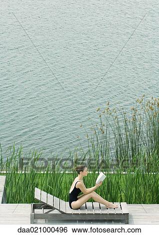 Stock Photograph - Woman sitting up on lounge chair, reading, next to lake. Fotosearch
