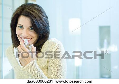 Stock Image - Woman smelling perfume bottle, smiling at camera. Fotosearch