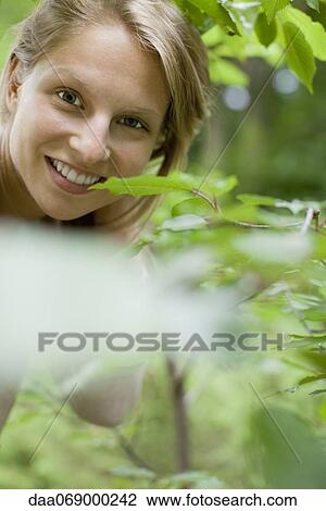Stock Image - Woman smiling at camera from behind foliage, portrait. Fotosearch