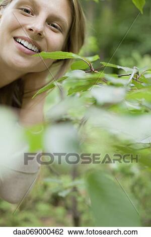 Stock Image - Woman smiling at camera from behind foliage, portrait. Fotosearch