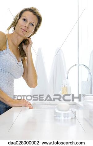 Stock Image - Woman standing by bathroom sink, smiling at camera. Fotosearch