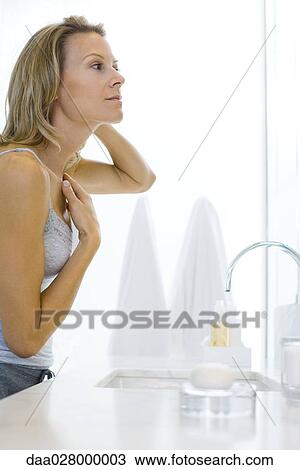 Stock Image - Woman standing by bathroom sink, examining self in mirror. Fotosearch