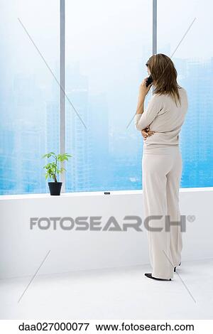 Woman standing by window, using cell phone View Large Photo Image Stock Photo - Woman standing by window, using cell phone. Fotosearch