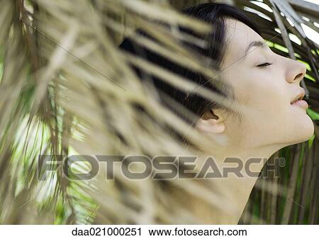 Stock Image - Woman standing in foliage, eyes closed, side view. Fotosearch