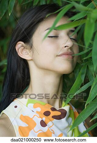 Stock Image - Woman standing in lush foliage, eyes closed, smiling. Fotosearch