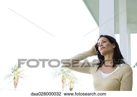Stock Image - Woman standing on porch, pushing hair back, looking into distance, palm trees in background. Fotosearch