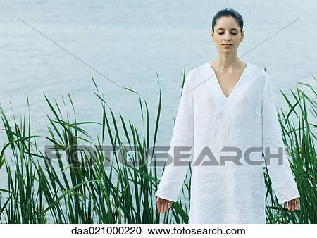 Stock Image - Woman standing with arms out near water's edge, eyes closed. Fotosearch