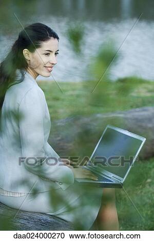 Stock Image - Woman using laptop in park, lake in background. Fotosearch