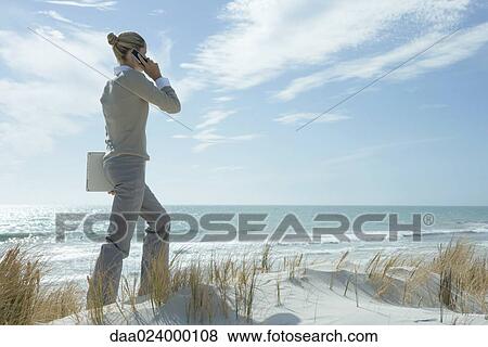 Stock Photo - Woman walking across dunes, using cell phone, carrying laptop under arm. Fotosearch