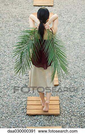 Stock Image - Woman walking on footpath, holding palm leaves behind back. Fotosearch
