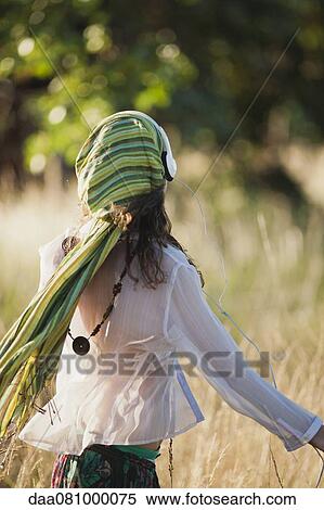 Stock Photography - Woman walking through field, listening to headphones, rear view. Fotosearch