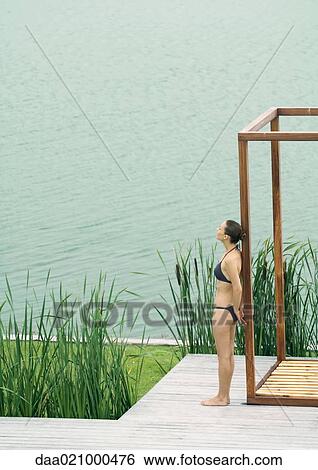 Stock Photograph - Woman wearing bikini, leaning against wooden structure, near lake. Fotosearch