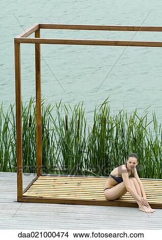 Picture - Woman wearing bikini, sitting in wooden structure, near lake. Fotosearch