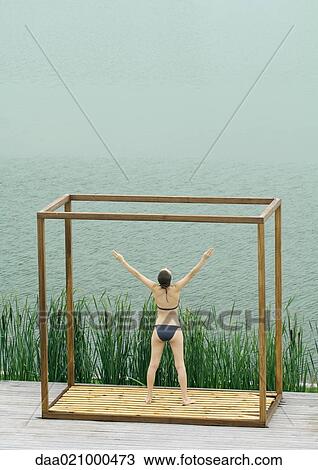 Woman wearing bikini, standing in square structure near lake, arms raised, rear view View Large Photo Image Stock Image - Woman wearing bikini, standing in square structure near lake, arms raised, rear view. Fotosearch