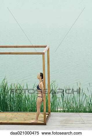 Stock Photography - Woman wearing bikini, standing in wooden structure, near lake. Fotosearch