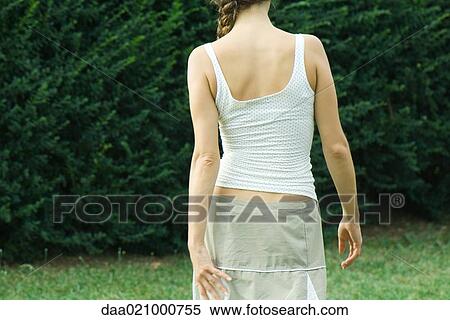 Stock Photography - Woman wearing skirt and tank top, walking toward hedge, cropped, rear view. Fotosearch