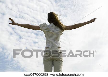Stock Photo - Woman with arms outstretched, cloudy sky in background, rear view. Fotosearch
