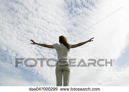 Stock Photo - Woman with arms outstretched, cloudy sky in background, rear view. Fotosearch