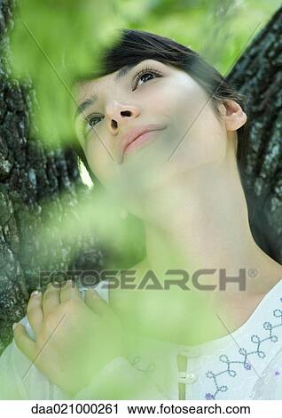 Stock Image - Woman with head against tree trunk, looking up. Fotosearch