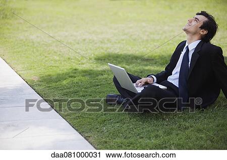 Young businessman sitting on grass with laptop computer, looking up and smiling View Large Photo Image Stock Image - Young businessman sitting on grass with laptop computer, looking up and smiling. Fotosearch