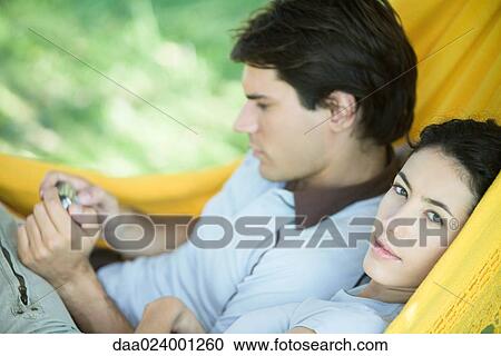 Stock Image - Young couple lying in hammock, using digital camera, woman looking at camera. Fotosearch