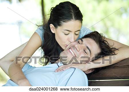 Stock Photo - Young couple, man sitting with head back while woman puts kisses his cheek. Fotosearch