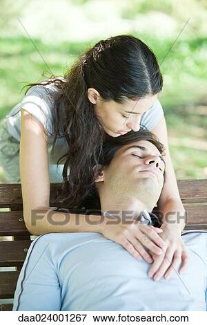Stock Photo - Young couple, man sleeping on bench while woman puts arms around him from behind. Fotosearch