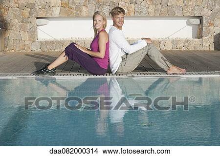 Picture - Young couple sitting back to back at poolside, portrait. Fotosearch