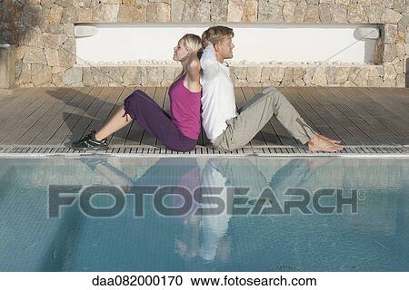 Stock Image - Young couple sitting back to back at poolside, hands behind heads. Fotosearch