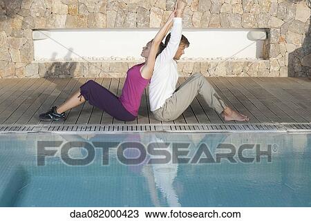 Stock Image - Young couple sitting back to back at poolside stretching. Fotosearch