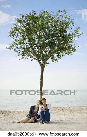 Stock Photo - Young couple sitting together beneath tree on beach. Fotosearch