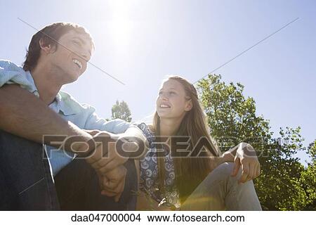 Young couple sitting together outdoors on sunny day, chatting, low angle view View Large Photo Image Picture - Young couple sitting together outdoors on sunny day, chatting, low angle view. Fotosearch