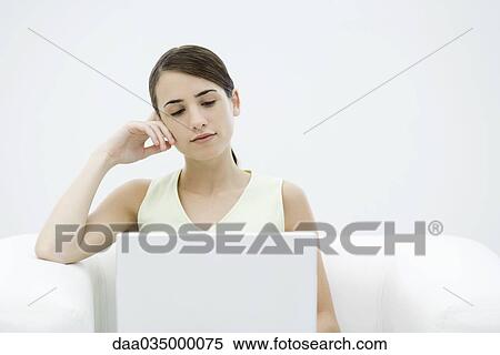 Stock Photography - Young female sitting in armchair, leaning on elbow, looking down at laptop computer. Fotosearch