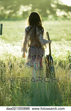 Stock Photograph - Young hippie woman holding guitar, standing in field, rear view. Fotosearch