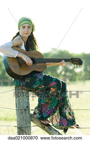 Stock Image - Young hippie woman sitting on fence post, playing guitar. Fotosearch