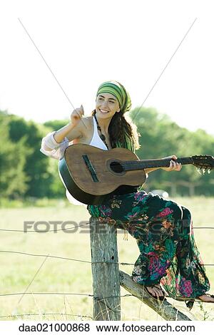 Stock Photo - Young hippie woman sitting on fence post, playing guitar. Fotosearch