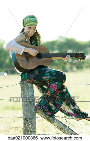 Stock Photograph - Young hippie woman sitting on fence post, playing guitar. Fotosearch