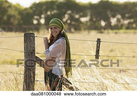 Young hippie woman standing by fence in field View Large Photo Image Stock Photo - Young hippie woman standing by fence in field. Fotosearch