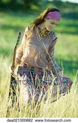 Stock Image - Young hippie woman walking through field, holding guitar. Fotosearch
