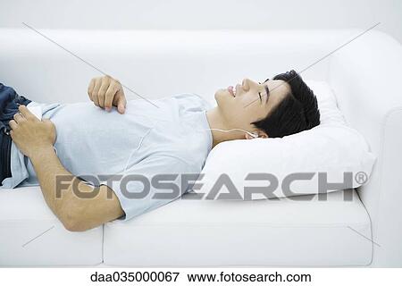 Young male listening to earphones, lying on couch, eyes closed View Large Photo Image Stock Photo - Young male listening to earphones, lying on couch, eyes closed. Fotosearch