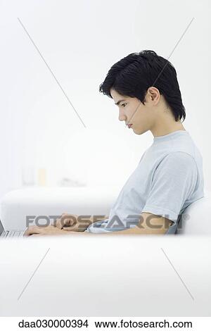 Young male sitting on sofa, using laptop, side view View Large Photo Image Picture - Young male sitting on sofa, using laptop, side view. Fotosearch