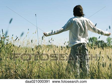 Stock Image - Young male standing in field, arms out, rear view. Fotosearch