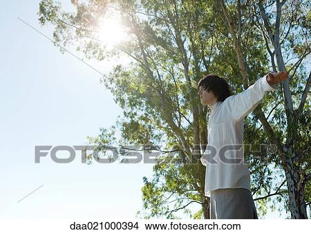 Young male standing outdoors, arms out, trees and sun in background View Large Photo Image Picture - Young male standing outdoors, arms out, trees and sun in background. Fotosearch