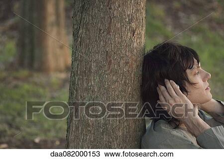Stock Image - Young man leaning against tree trunk, listening to music with headphones, side view. Fotosearch