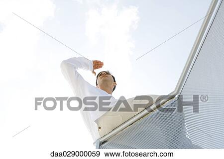 Stock Photo - Young man leaning on rail, shading eyes, looking into distance, low angle view. Fotosearch