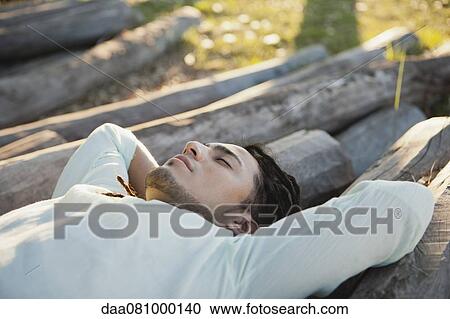 Stock Image - Young man lying on pile of logs, eyes closed. Fotosearch
