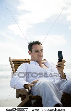 Young man sitting in chair on beach, using cell phone View Large Photo Image Stock Image - Young man sitting in chair on beach, using cell phone. Fotosearch