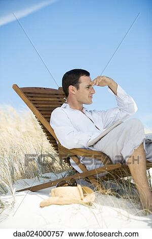 Young man sitting in deck chair on beach, shading eyes View Large Photo Image Stock Photo - Young man sitting in deck chair on beach, shading eyes. Fotosearch
